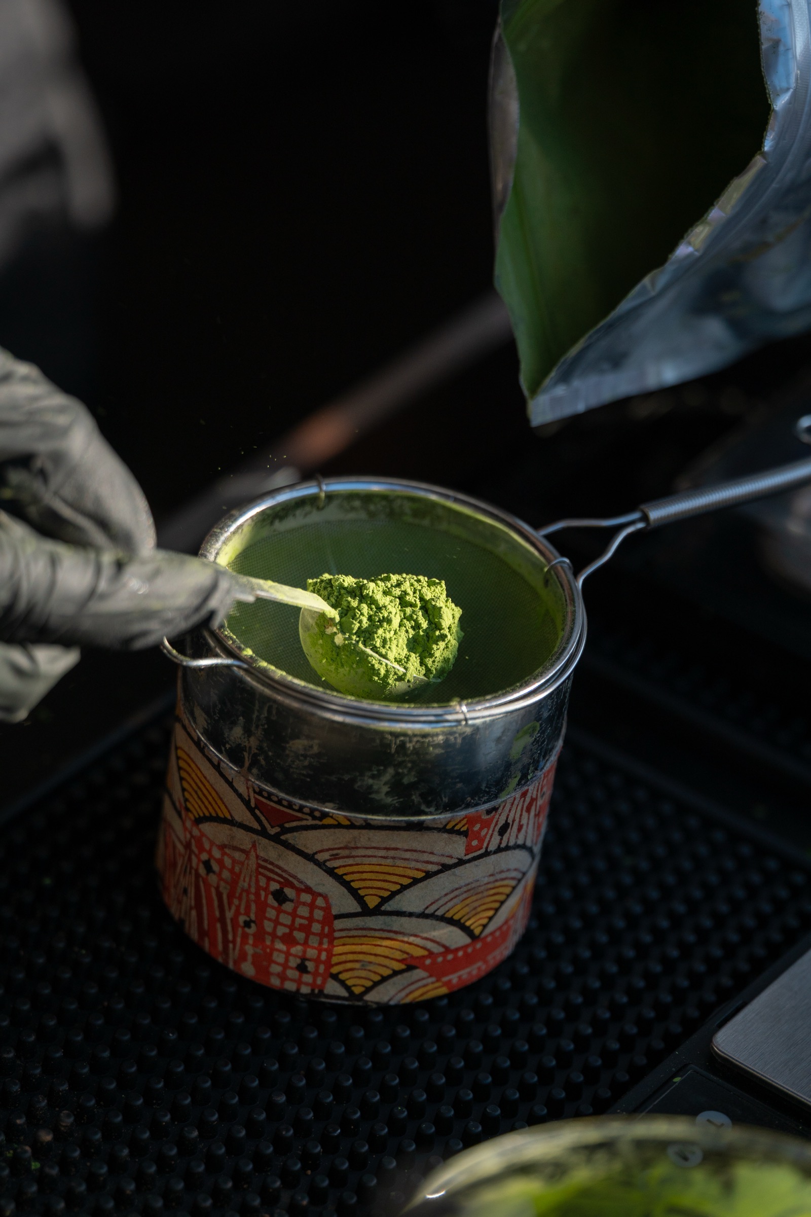 A gloved hand sifting bright-green ceremonial matcha through a strainer into a patterned tin
